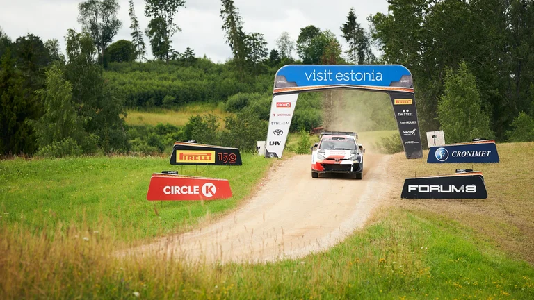 A rally car speeding on a dirt track surrounded by lush green fields, under an archway that reads "Visit Estonia," with sponsor banners on both sides of the track.