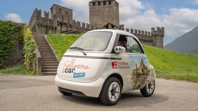 A small car with two people inside. On the backgroud, a medieval castle, an hill and a green meadow.