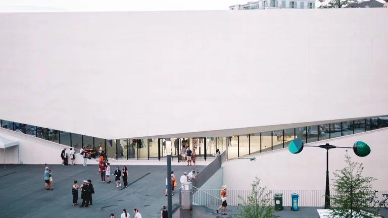 People gather outside a modern building with a large white roof.