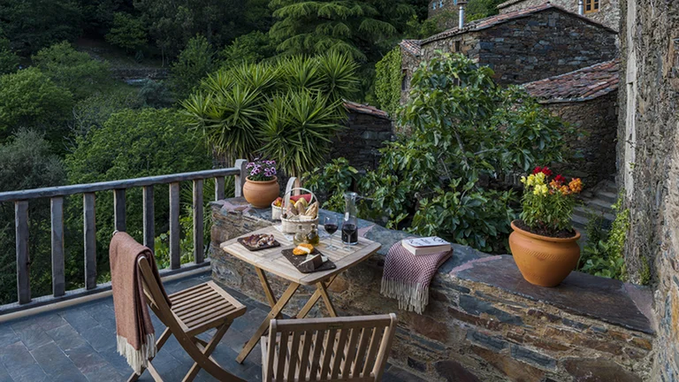 Balcony with table set for a meal, overlooking lush greenery and a stone building in Cerdeira, Portugal.