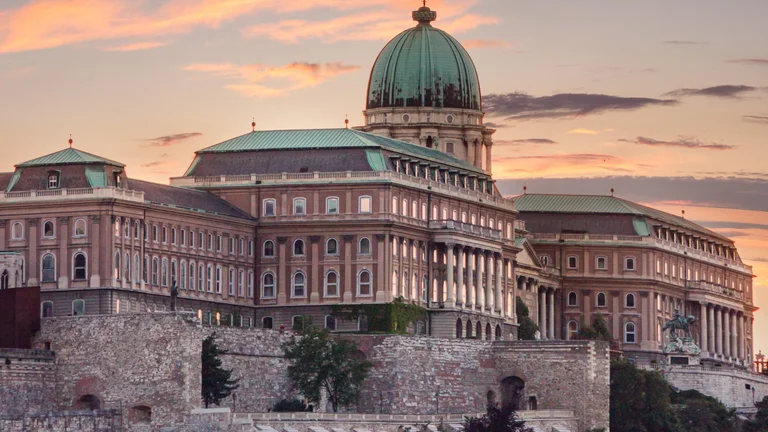 The Buda Castle in Budapest, Hungary, with its green dome and historic architecture illuminated against a dramatic orange and purple sunset sky.