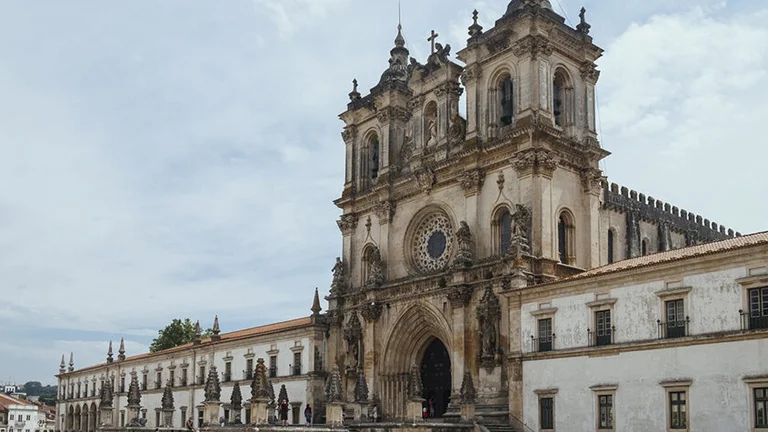 Ornate historical Monastery of Alcobaça with visitors at the entrance under a cloudy sky.