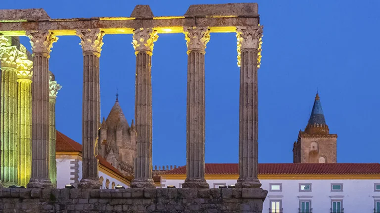Ancient Evora Roman temple columns lit at night with modern buildings and a church in the background.