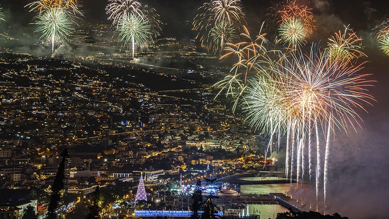 Fireworks display over the Madeira island at night with illuminated streets and a large Christmas tree.