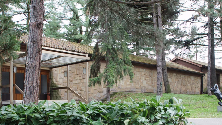 Stone building with large windows surrounded by pine trees and a sculpture of a reclining figure.