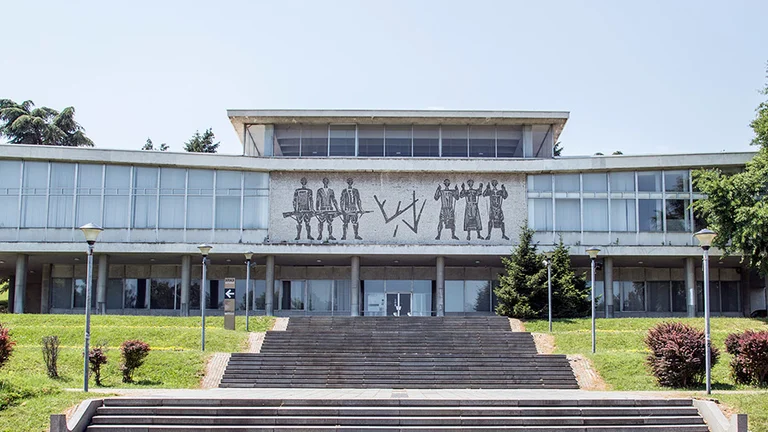 Museum of Yugoslavia is a building with a mural, stairs in front, surrounded by trees under a clear sky.
