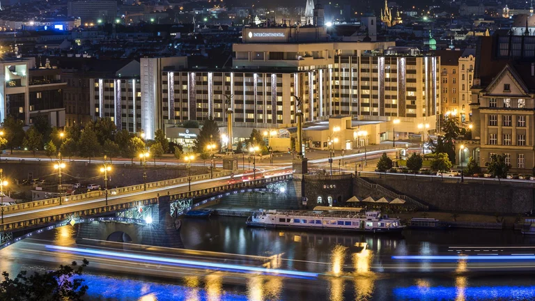 Nighttime cityscape of Prague featuring Intercontinental hotel, a bridge, and light trails from moving boats on the water.
