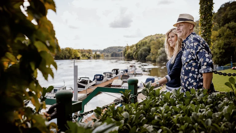 Couple standing by the Vltava river overlook with moored boats and lush greenery.