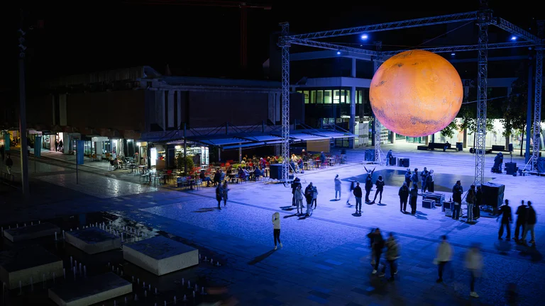 Nighttime view of an outdoor square with people and a large suspended glowing Mars replica.