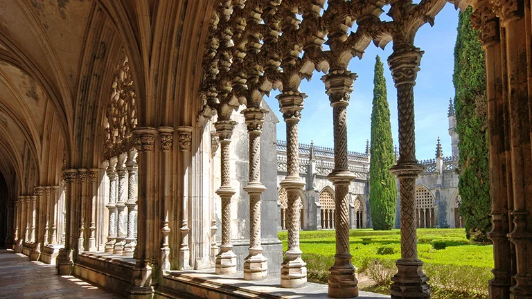 Gothic cloister with ornate pillars overlooking a green courtyard and clear sky at the Mosteiro Batalha in Portugal.