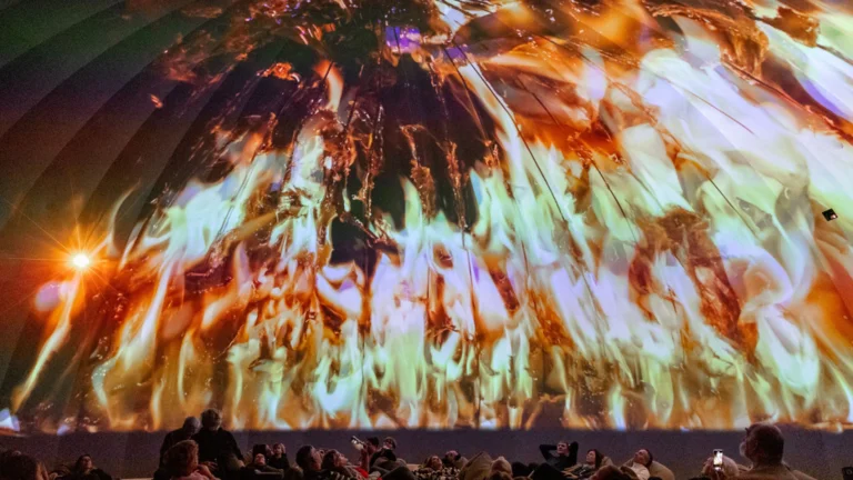 An audience watching a vibrant light projection on a dome ceiling.