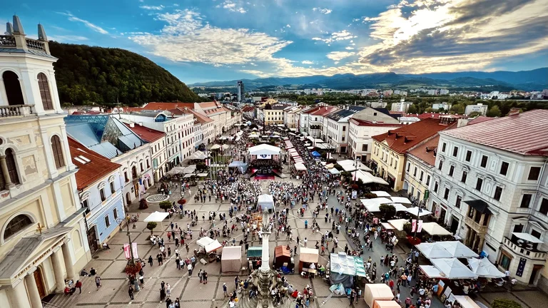 Aerial view of a Banská Bystrica square during the Radvanský fair, with tents and crowds, against a backdrop of mountains and a dramatic sky.