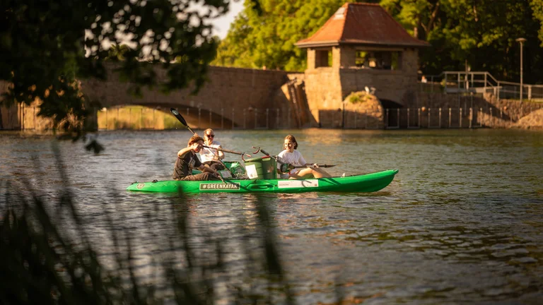 A group of three people kayaking along a river near a stone bridge and greenery.