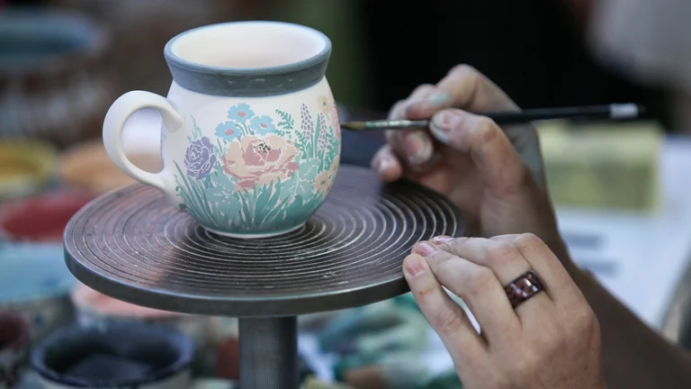 A potter carefully paints floral designs on a pottery mug, using a fine brush on a spinning turntable surrounded by colorful paints.