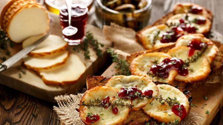 Rustic wooden table with toasted bread topped with melted cheese, cranberry sauce, and fresh thyme, alongside sliced cheese and condiments.