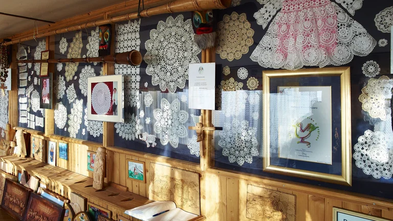 A wooden wall adorned with intricate lace artworks, a pink lace dress, framed pictures, and carved wooden decorations.