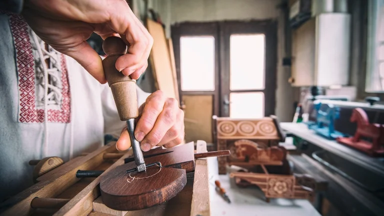 A craftsman carves wood with a chisel in a workshop, showcasing intricate details on the wood and traditional attire.