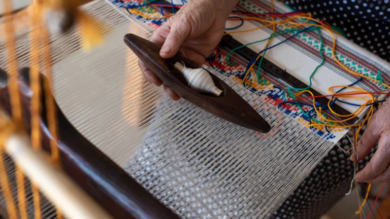 A close-up of a hand weaving on a loom, holding a shuttle with thread, colorful yarns interwoven, showcasing traditional craftsmanship.