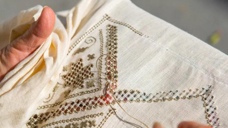 A close-up of a hand holding embroidered fabric, showcasing intricate stitching patterns and a sewing needle with thread.
