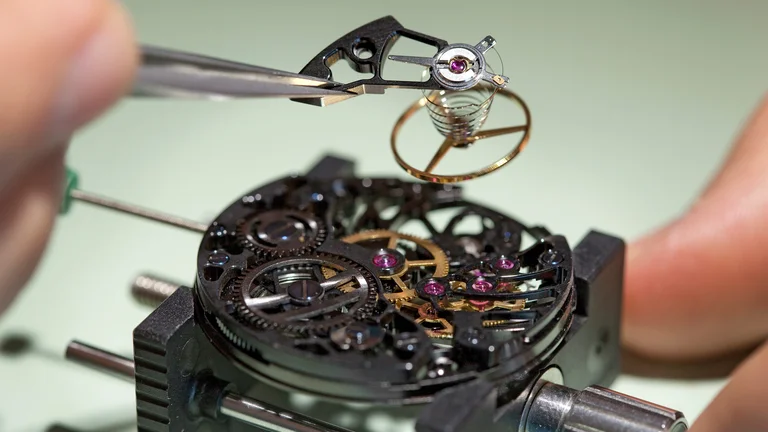 A close-up of a watchmaker using tweezers to manipulate a delicate escapement mechanism above a detailed watch movement with gears and jewels.
