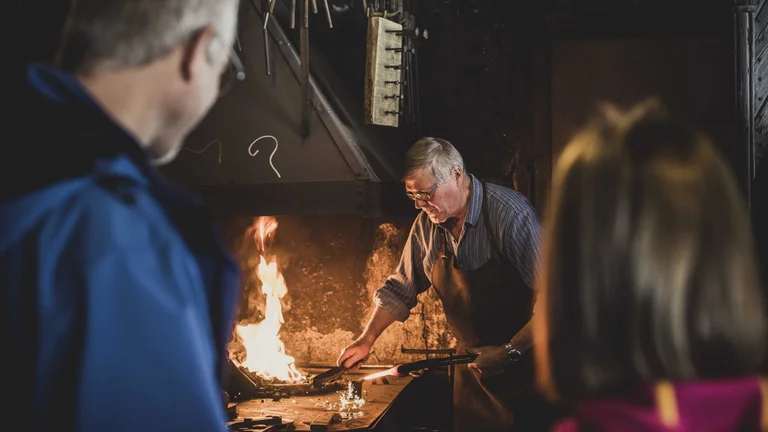 A blacksmith skillfully works over a glowing forge, demonstrating traditional metalworking techniques to intrigued observers.