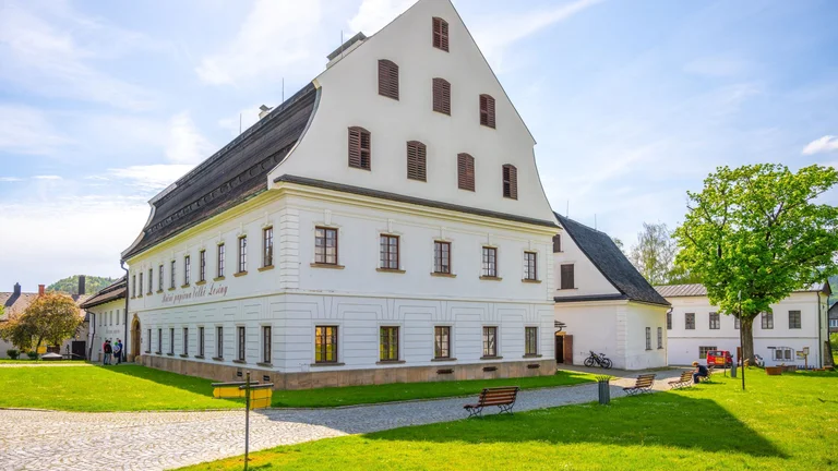A white historic building with sloped roofs and windows, surrounded by green grass, benches, and trees on a sunny day.