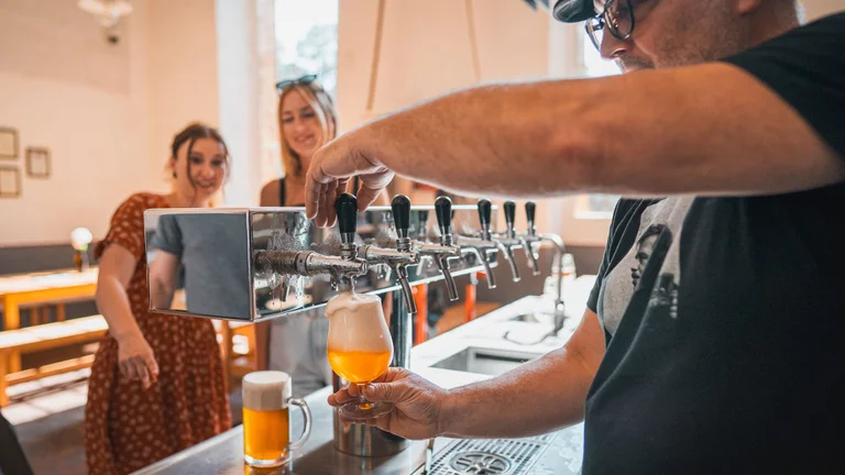 A bartender pours a frothy, golden beer into a glass, surrounded by patrons in a lively brewery setting.