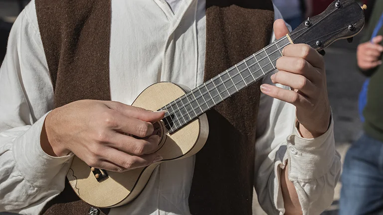 A close-up of a person in a white shirt and brown vest playing a small wooden ukulele, fingers poised on the frets.