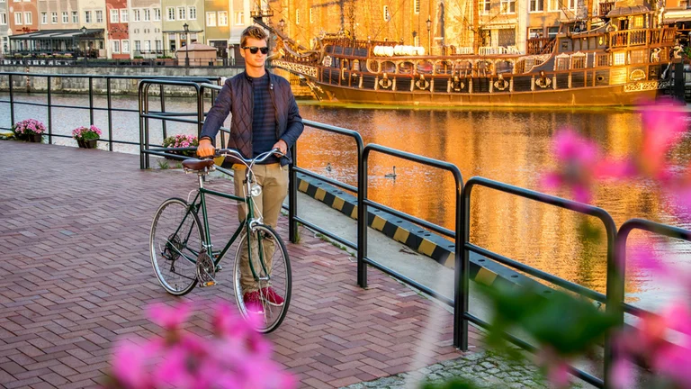 A person stands next to their bicycle by a canal in Gdańsk, surrounded by blooming flowers and historic buildings, with a ship in the background.