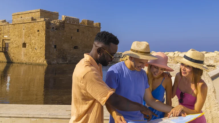 A group examines a map near a historic stone castle in Pafos by the sea on a sunny day, wearing summer hats.