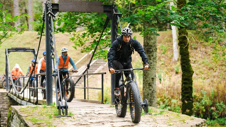A cyclist traverses a stone bridge surrounded by greenery, while others follow behind in bright orange vests.