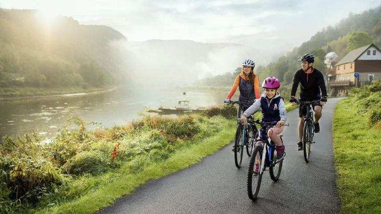 A family cycle along a the Elbe cycle route, along a misty riverbank, surrounded by lush greenery and rolling hills under the soft morning light.