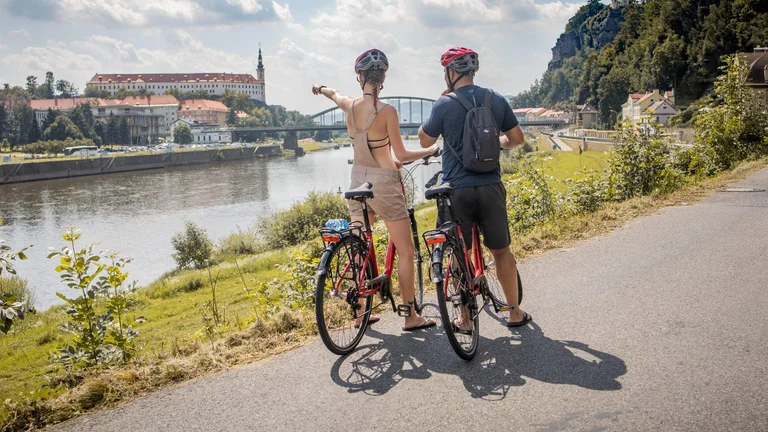 A couple on bicycles, wearing helmets, admires a river view and points at a nearby historic building under a sunny sky.