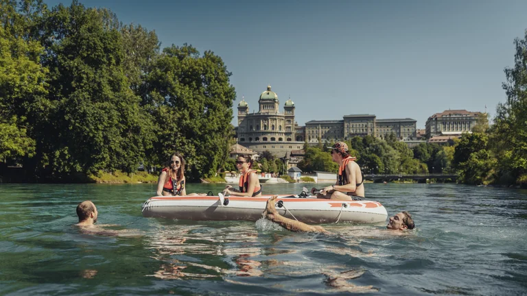 A group enjoys a sunny day on the water in Bern, with a boat drifting by lush greenery and a historic building in the background.