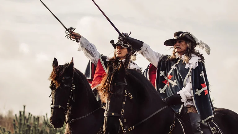 Two people in historical musketeer costumes raise swords while riding black horses during a reenactment.