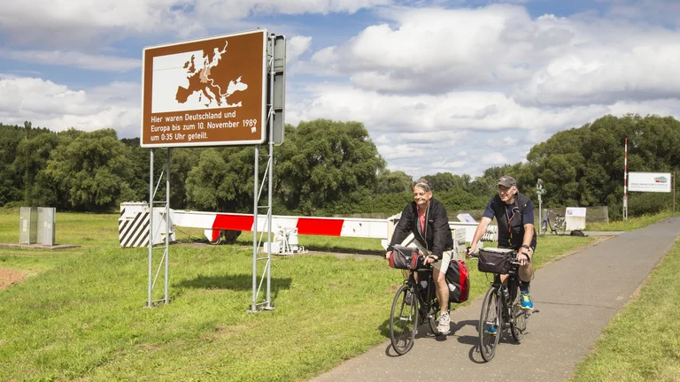 Two cyclists ride along a path with a sign marking the historical division of Germany and Europe until 10 November 1989.