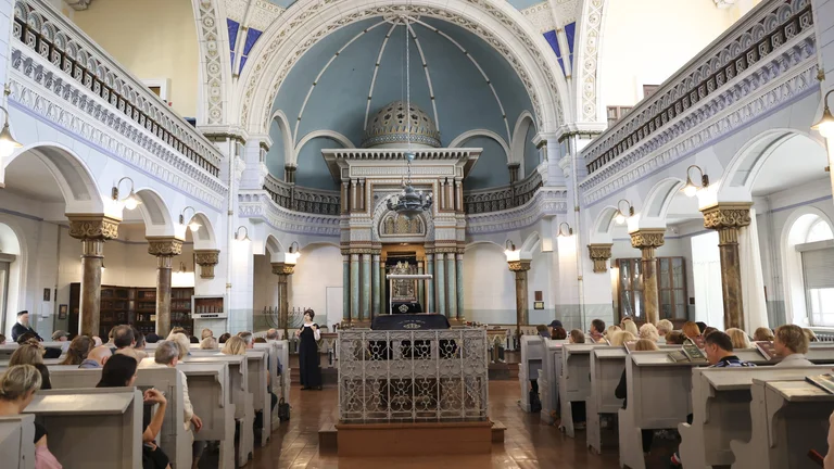 A spacious synagogue interior with decorative arches and domed ceiling, filled with seated visitors listening to a speaker.