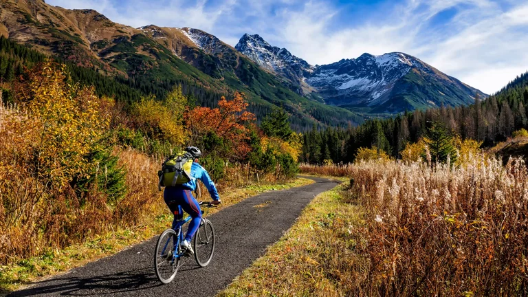 A cyclist biking on a mountain trail framed by autumn foliage and towering peaks, with the High Tatras in the background.