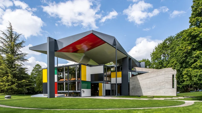 The Pavillon Le Corbusier in Zurich, a modernist architectural masterpiece featuring a geometric design with bold primary colors—red, yellow, blue, and green—contrasting against black and white panels. The structure is surrounded by lush green grass and trees, with a large angular roof extending outward, creating a striking visual effect. The sky is bright blue with scattered white clouds.