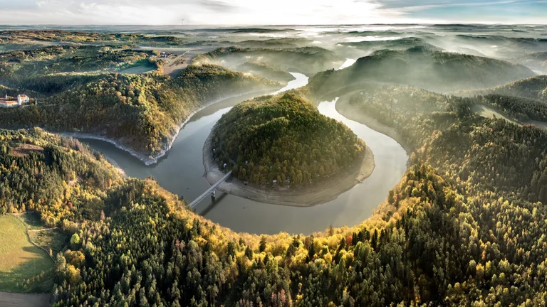 Aerial view of lush hills and winding river, with a historic castle nestled among colorful autumn foliage under a misty sky.