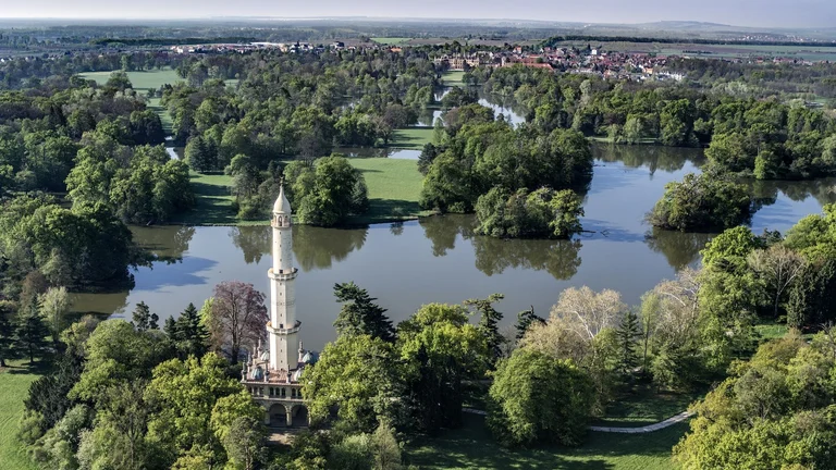 A scenic landscape featuring a tall white tower surrounded by lush greenery and a winding river, with a distant village in the background.