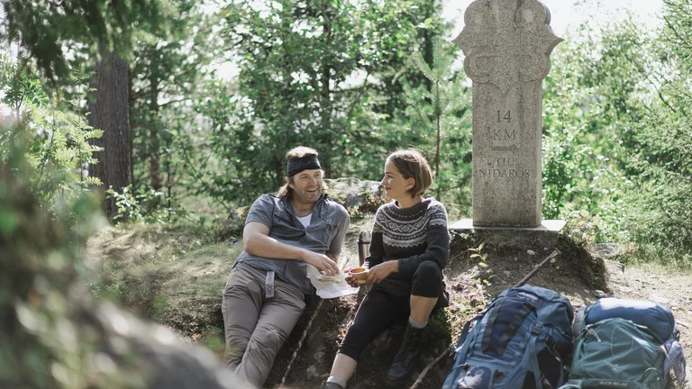 Two hikers resting by a historic stone marker on a forest trail, surrounded by lush greenery.