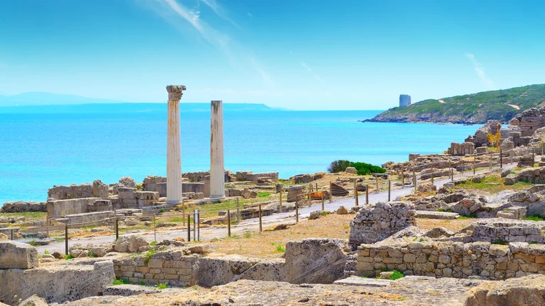 Ancient ruins with columns overlooking tranquil blue waters, set against a clear sky and distant green hills.