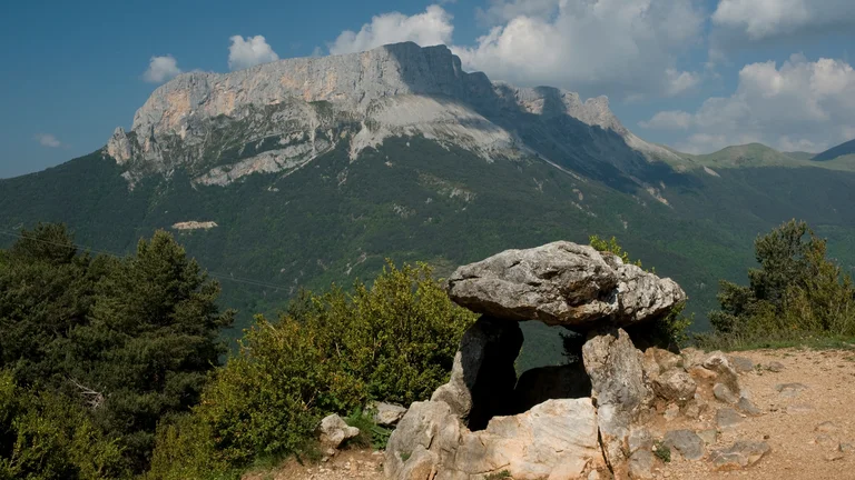 A stone dolmen stands in the foreground, framed by lush greenery and a backdrop of rugged mountains under a blue sky with scattered clouds.