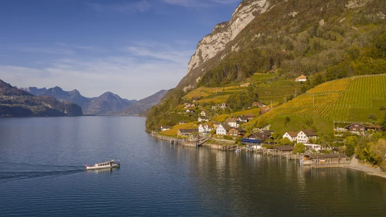 A peaceful lakeside village with terraced vineyards nestled against steep mountains, with a boat gliding across calm blue waters.