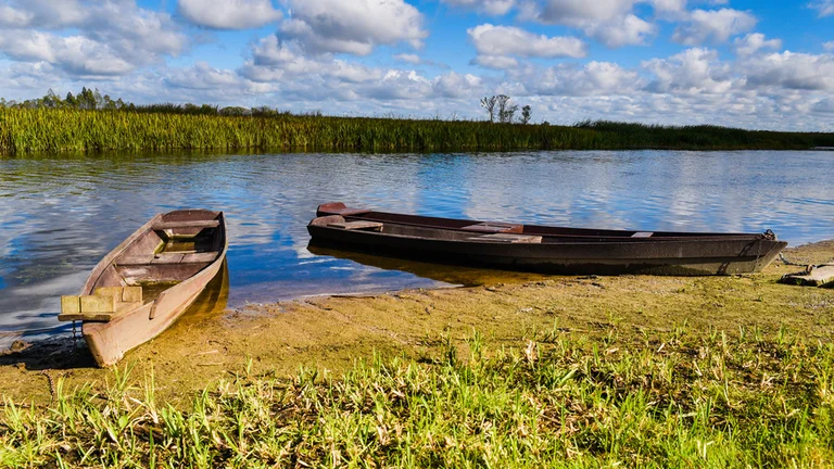 Two wooden boats rest on a sandy shore beside a calm river, surrounded by lush green grass and under a partly cloudy sky.