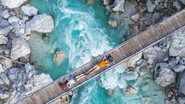 Aerial view of a wooden bridge over a turquoise river surrounded by rocky banks, with a person lying on the bridge.