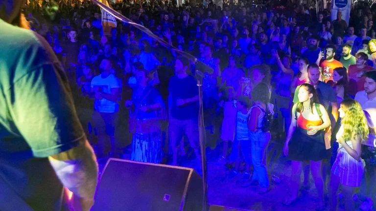 A crowded outdoor music festival at night, illuminated by colorful lights, featuring a performer in the foreground.