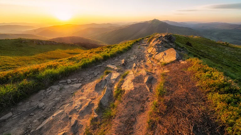 A rocky hiking trail winds along a hillside at sunset, with golden light illuminating the mountains in the background.