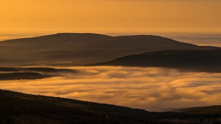Golden-hued mountain range shrouded in mist, with layers of clouds and distant peaks under a soft sunset glow.